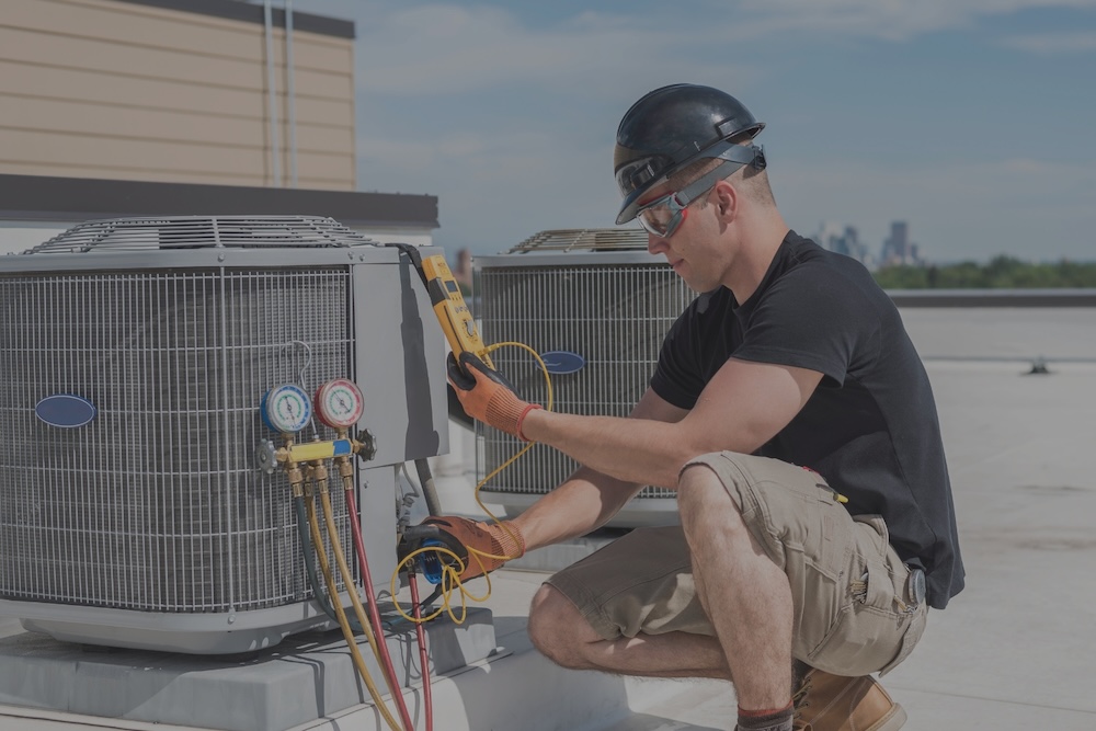 An HVAC technician wearing safety gear crouches on a rooftop to inspect an outdoor air conditioner unit using diagnostic tools.