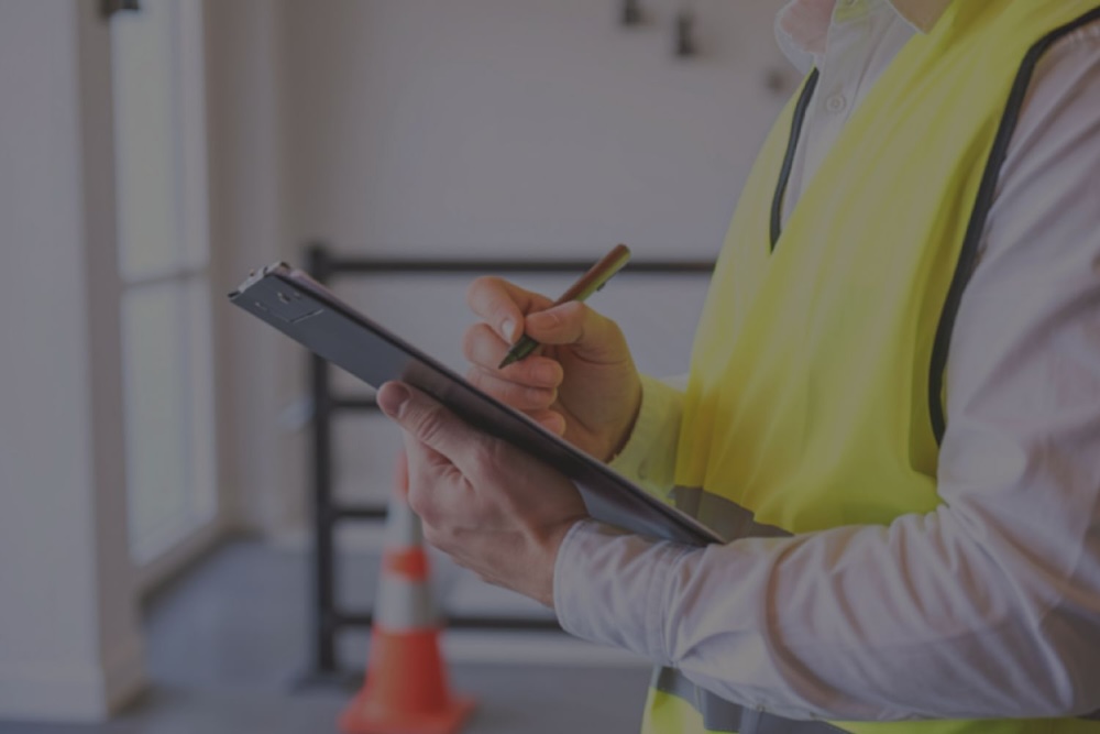 An environmental professional wearing a safety vest takes notes on a clipboard during a commercial property site inspection.